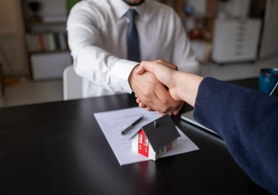 Two people shake hands across a desk with a contract, pen, and small model house, suggesting a real estate or property agreement.