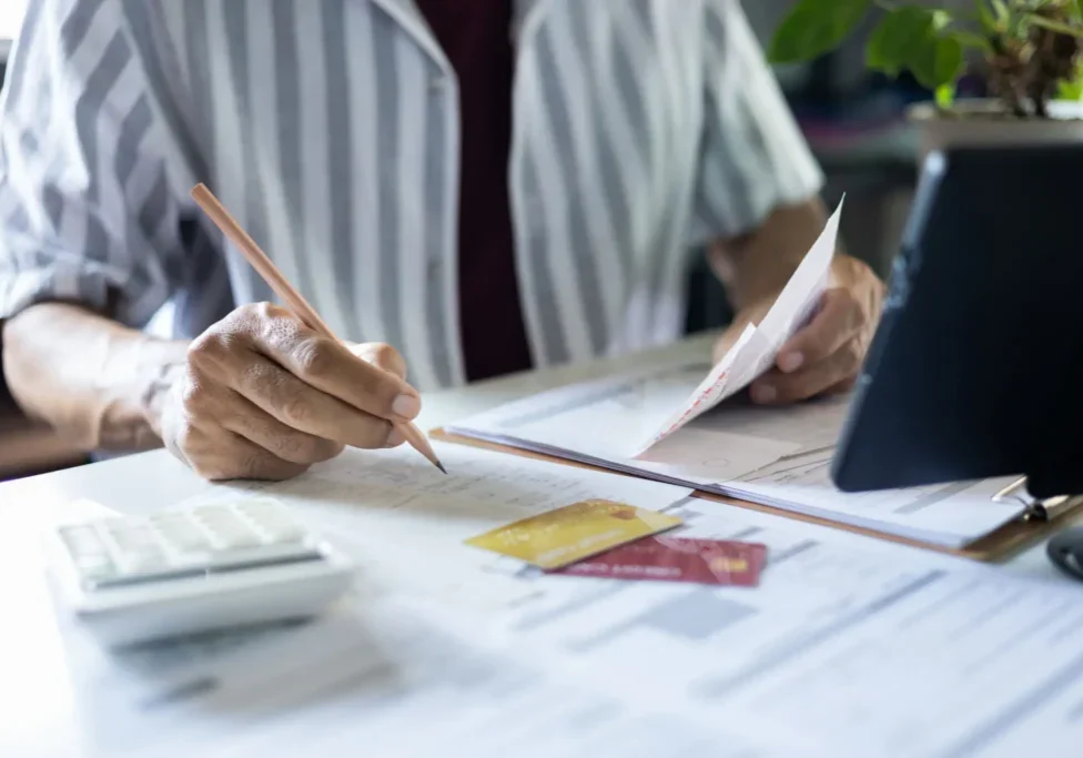Person in a striped shirt writes on financial documents at a desk, with calculator, credit cards, papers, and tablet nearby; focused office setting.